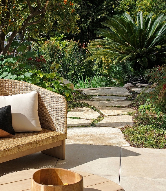 Canapé en osier sur terrasse pavée menant à un chemin de pierres dans un jardin luxuriant avec oranger et palmier.